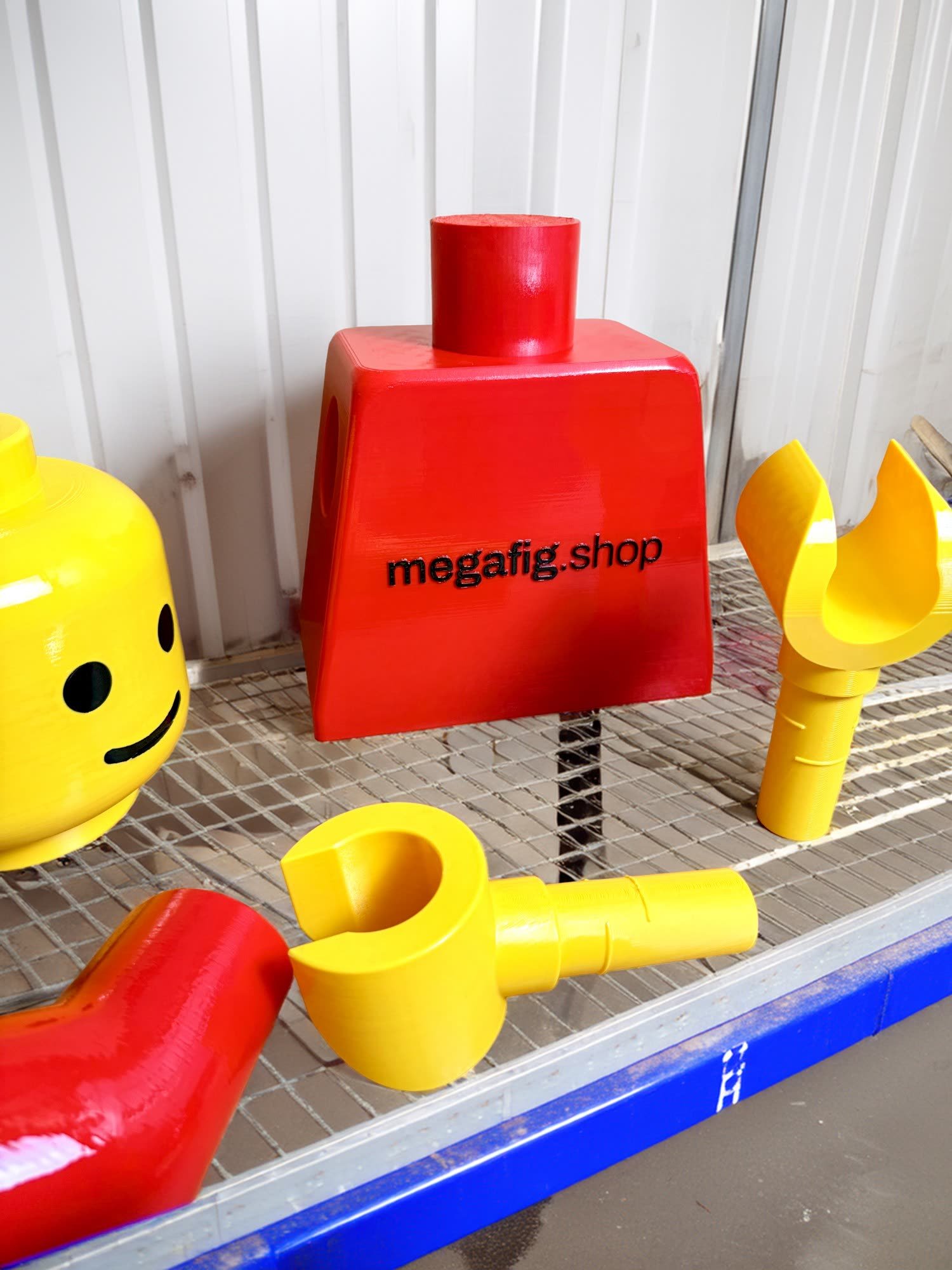 Freshly clear-coated Mega Figure parts on a drying rack in the studio spray booth — a yellow head, a red torso embossed megafig.shop, and yellow hands, all in high gloss