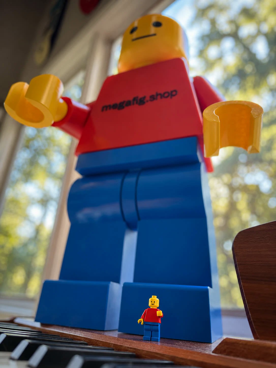 A 24-inch gloss Mega Figure standing on a piano lid beside a standard minifigure, shot from below against a sunlit window