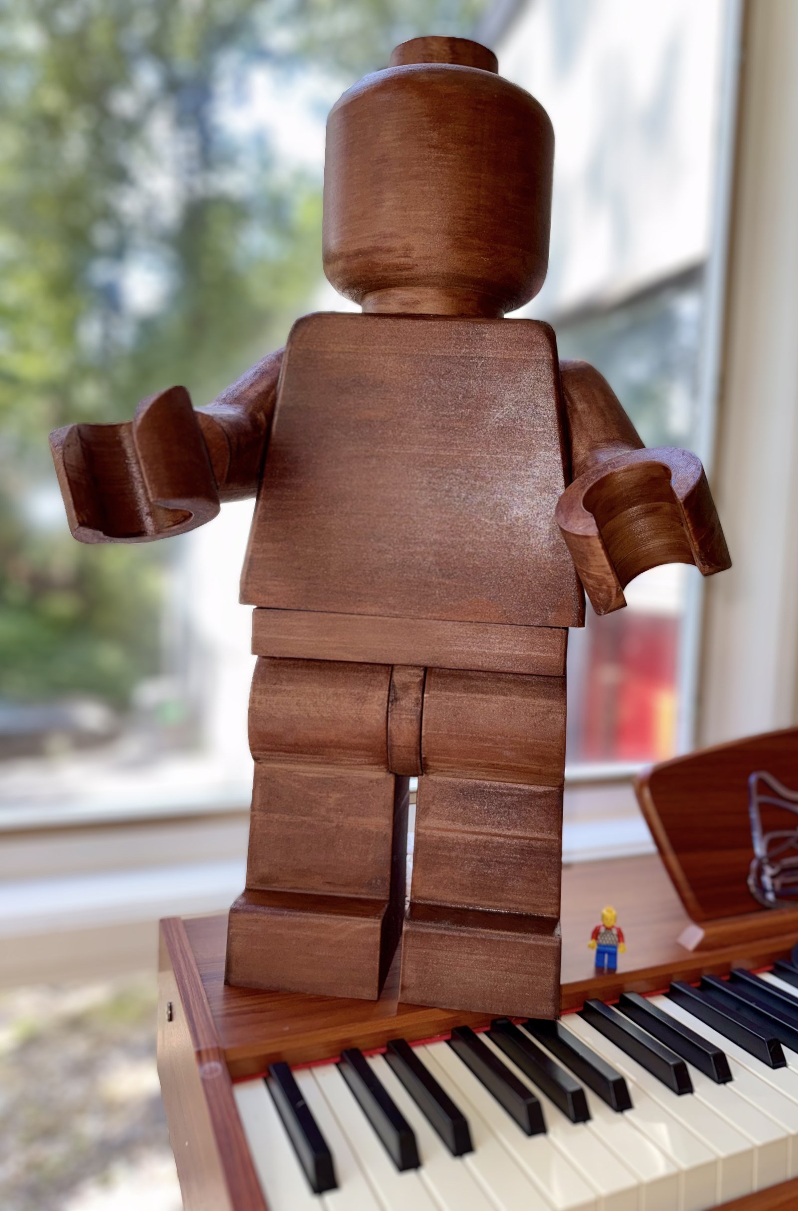 The Wood Edition standing on a piano beside a standard minifigure, showing how much larger it is