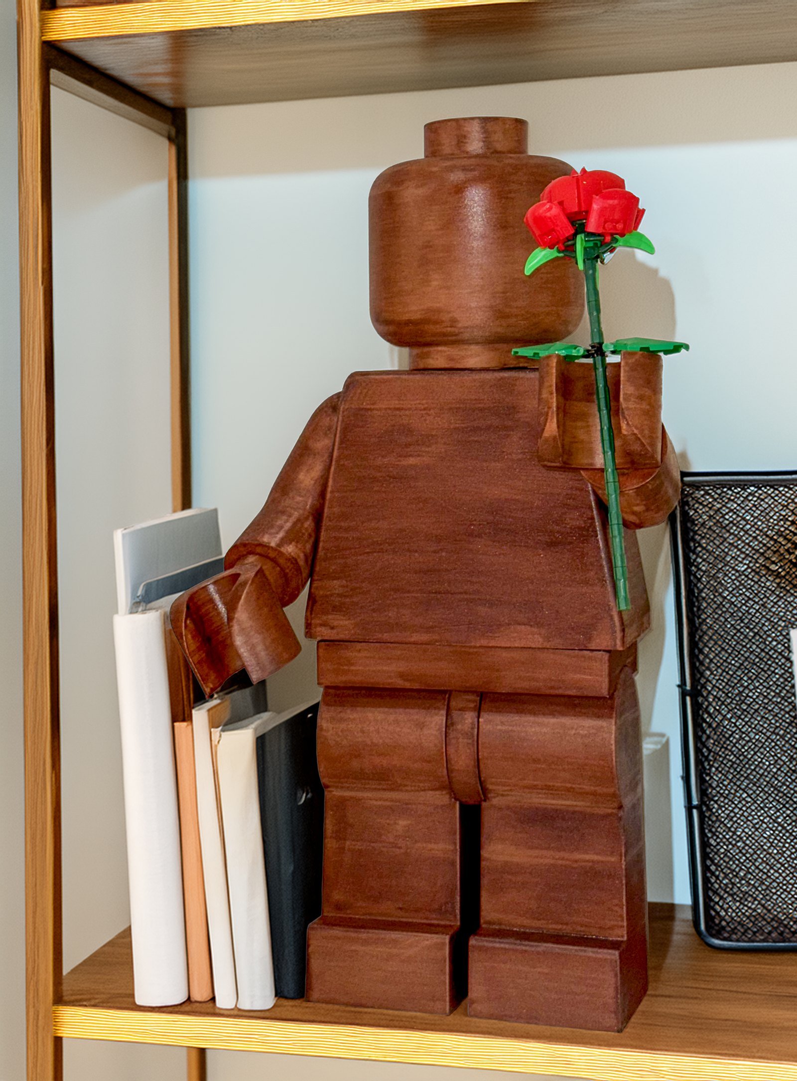 The Wood Edition figure on a warmly lit bookshelf beside a row of books, holding a single red rose